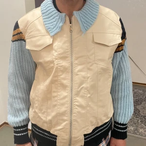 Beige jacka med stickade ärmar från Fashion - Unik beige jacka med stickade ljusblå ärmar och krage. Jackan har dragkedja framtill, två bröstfickor med lock och svarta detaljer på ärmarna. Materialmix med syntet och stickat ger en cool look. Storlek S/M