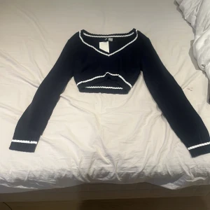 Mörkblå croppad stickad tröja från H&M Divided - Säljer en mörkblå croppad stickad tröja från H&M Divided med vita ränder längs v-ringningen, ärmsluten och nederkanten. Tröjan har lång ärm och en tight passform, perfekt för att skapa en trendig look. Endast använd vid spegel bilden.