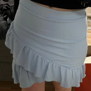 Ljusblå volangkjol från NellybyNelly - Supersöt ljusblå kjol från Nelly med volangdetalj längs nederkanten. Kjolen är kort och har en figurnära passform som framhäver siluetten. Perfekt för vår och sommar, tillverkad i ett lätt och bekvämt material.