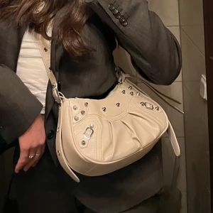 Beige väska med nitar och silverdetaljer - Säljer en snygg beige axelväska med silverfärgade metalldetaljer och dragkedja. Väskan har en justerbar axelrem med flätad detalj och cool design med nitar och spännen. Perfekt för att lyfta vilken outfit som helst!