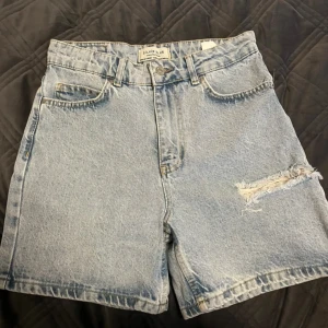 Ljusblå jeansshorts från Dilvin Blue - Säljer ett par ljusblå jeansshorts från Dilvin Blue med klassisk femficksdesign och slitning på ena benet. Shorts med hög/mellan midja och råa kanter, perfekt för en avslappnad stil.