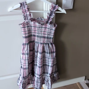 Rutig rosa och svart klänning med volang - Supersöt kort klänning från NA-KD i rosa, svart och vit rutig bomull. Smockad överdel, volangdetaljer på axelband och nederkant. Perfekt för sommarens varma dagar!