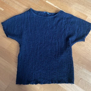 Mörkblå strukturerad topp från 157 - Säljer en mörkblå topp från 157 i storlek XS. Toppen har en unik strukturerad, nästan krinklad yta och korta ärmar. Den har en rak passform och rund halsringning. Perfekt för att ge en cool touch till din outfit!