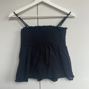 Svart smockad linnetopp från Gina Tricot - Säljer en svart smockad linnetopp från Gina Tricot med tunna axelband och volangkant upptill. Toppen har en luftig och lätt passform, perfekt för varma dagar. Enkel och stilren design som passar till mycket. Skulle säga att den har grymt lite.