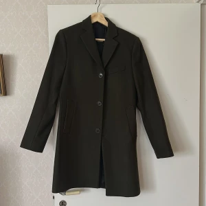 Olivgrön rock från J.Lindeberg - Snygg olivgrön rock från J.Lindeberg med klassisk krage och tre knappar framtill. Jackan har långa ärmar med fyra dekorativa knappar vid ärmsluten och diskreta fickor på sidorna. Perfekt för dig som vill ha en stilren och tidlös look. Slimfit. Användes ej då den var något för liten.