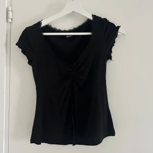 Svart topp med spets från Gina Tricot - Säljer en svart topp från Gina Tricot i storlek XS. Toppen har v-ringning med spetsdetaljer vid halsen och korta ärmar. Den har en lätt rynkning framtill som ger en snygg passform. Perfekt till många olika outfits!