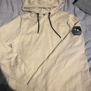 Beige hoodie från Jack & Jones Core - Säljer en beige hoodie från Jack & Jones Core med svart patch på ärmen och svarta dragsnören. Tröjan har lång ärm och huva, perfekt för en avslappnad stil.hör av er vid frågor och pris!