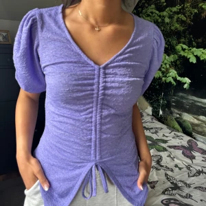 Ljuslila ribbad topp med puffärm och knyt - Superfin ljuslila topp med kort puffärm och v-ringning. Toppen har ribbad struktur och snygg rynkdetalj framtill med knyt i midjan. Köpt från BIKBOK i storlek S.