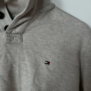 Beige långärmad tröja från Tommy Hilfiger - Snygg beige långärmad tröja från Tommy Hilfiger med hög krage och knappar. Tröjan har ett diskret broderat logotyp på bröstet och ribbade muddar. Perfekt för en stilren och avslappnad look.