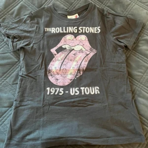 Grå Rolling Stones t-shirt från H&M - Säljer en grå t-shirt från H&M med Rolling Stones tryck och ikoniska tungan på framsidan. T-shirten har korta ärmar och är i mjuk bomull. Perfekt för dig som gillar bandtröjor och vill ha en cool vintage-look.