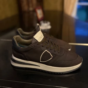 Bruna/Olivgröna sneakers från Philippe Model - Säljer ett par bruna/olivgröna sneakers från Philippe Model med vit sula och svarta detaljer. Skorna har snörning och en ikonisk sköld-logga på sidan. Perfekta för dig som gillar stilrena och bekväma sneakers.