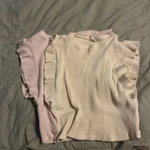 Ribbade toppar med volangärmar - Två ribbade toppar i ljusrosa och offwhite med volangdetaljer längs ärmarna. Topp med rund halsringning och korta ärmar, perfekt för en söt och stilren look. Materialet är mjukt och stretchigt.