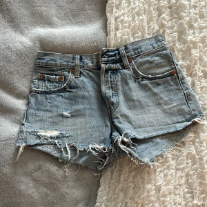 Ljusblå jeansshorts från Levi's - Säljer ett par ljusblå jeansshorts från Levi's med fransig kant och slitna detaljer. Klassisk femficksmodell med hög midja och råa benslut. Perfekta för sommaren och ger en avslappnad look.