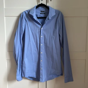 Ljusblå skjorta från Tiger of Sweden - Säljer en klassisk ljusblå skjorta från Tiger of Sweden med slim fit-passform. Skjortan har långa ärmar, vit knappstängning och en stilren krage. Perfekt för dig som gillar en clean och tidlös look.