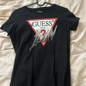 Svart t-shirt från Guess med strassdetaljer - Svart t-shirt från Guess med klassisk röd och vit logga framtill och glittrig strassdekor över bröstet. T-shirten har rund hals och korta ärmar.