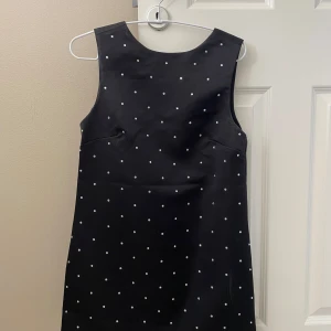 Svart prickig klänning från H&M - Säljer en svart ärmlös klänning från H&M med små diamanter och en söt rosett i ryggen. Klänningen har en djup v-ringning bak och är i en klassisk, rak modell. Perfekt för dig som gillar stilrena detaljer!