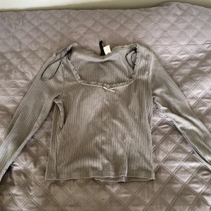 Grå ribbad topp från H&M Divided - Säljer en grå ribbad långärmad topp från H&M Divided. Tröjan har en fyrkantig ringning med spetskant och en liten rosett framtill. Säljer en likadan i vit