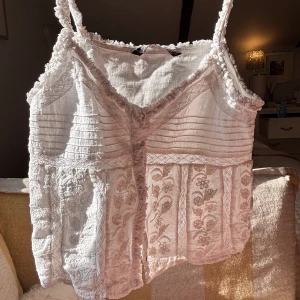 Vit linnetopp med spetsdetaljer från Zara - Säljer en söt vit linnetopp från Zara med smala axelband och fina spetsdetaljer. Toppen har v-ringning, dekorativa knappar framtill och broderade blommor nedtill. Perfekt för varma dagar och enkel att matcha. Endast använd ett fåtal gånger! Köpt för nått år sen så den säljs ej längre! Jätte nice att ha på stranden osv…🌸