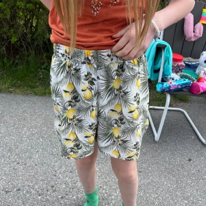 Vita shorts med citronmönster från Name It - Säljer ett par vita shorts från Name It med citroner och gröna blad som mönster. De har normal passform, resår i midjan och är perfekta för sommaren. Superfräscha och lätta att matcha med en enkel topp!