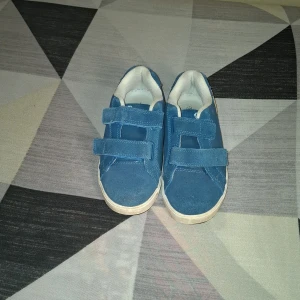 Blå sneakers med kardborreband - Säljer ett par blå sneakers från Champion med vita detaljer och två praktiska kardborreband. Skorna har vit sula och en gul/vit logga på sidan. Perfekta för dig som vill ha ett par snygga och bekväma vardagsskor.