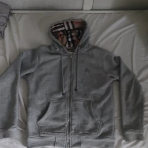 Grå hoodie från Burberry med dragkedja - Säljer en grå hoodie från Burberry med klassiskt rutigt foder i huvan och dragkedja framtill. Tröjan har två fickor och broderad logga på bröstet. Perfekt för dig som gillar stilrena och exklusiva plagg.