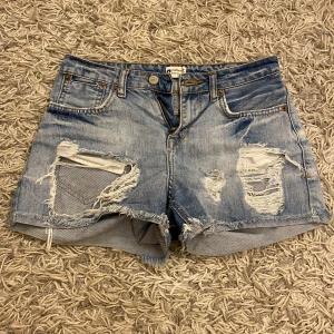Ljusa jeansshorts från Gina Tricot - Säljer ett par ljusblå jeansshorts från Gina Tricot med slitna detaljer och fransig kant. Shortsen har klassisk femficksmodell, knapp och dragkedja i gylfen samt mid till low waist. Perfekta för sommaren!