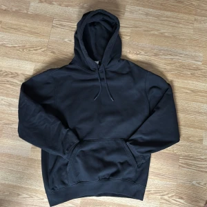 Svart hoodie från H&M Essentials - En klassisk svart hoodie från H&M Essentials med huva och känguruficka framtill. Modellen är relaxed fit och har ribbade muddar vid ärmslut och nederkant. Perfekt för en avslappnad stil. Stor i storleken