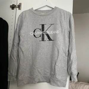 Grå sweatshirt från Calvin Klein Jeans - Säljer en ljusgrå sweatshirt från Calvin Klein Jeans med stor svart och vit logga på bröstet. Tröjan har rund halsringning och långa ärmar. Perfekt för en avslappnad och stilren look. Köpte den från designonly för 1000kr. Pris kan diskuteras vid snabb affär!!!