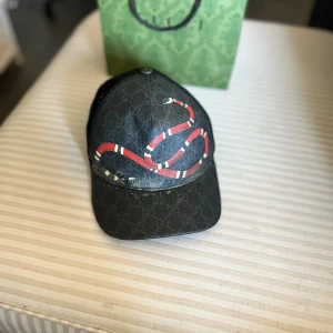 Svart keps från Gucci med orm-motiv - Säljer en svart keps från Gucci med ikoniskt orm-motiv framtill och diskret GG-mönster i bakgrunden. Kepsen har justerbar rem baktill med röd-blå detalj och mesh-paneler på sidorna för extra ventilation.