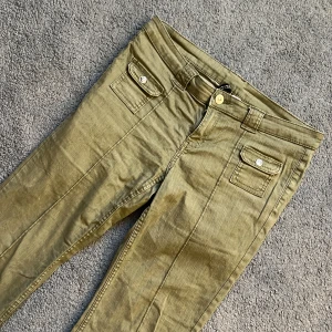 Olivgröna byxor med fickdetaljer från H&M Divided - Snygga olivgröna byxor från H&M Divided med raka ben och två fickor framtill med lock och knapp. Byxorna har normal passform och stängs med dragkedja och knapp i midjan. Perfekta för en avslappnad och trendig stil. De har inga bakfickor. Jag säljer dessa för att dem är för korta och stora för mig. de är knappt använda och har bara legat i min garderob (därav ser dem skrynkliga ut). Jag säljer dem för 500 kr + frakt!