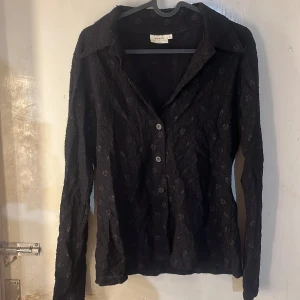  spetsskjorta - Säljer en svart skjorta från H&M Woman Collection med lång ärm och knappar framtill. Skjortan har ett diskret spetsmönster som ger en snygg textur och klassisk krage. Perfekt för dig som gillar stilrena plagg med en twist.