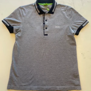 Grå piké t-shirt från Hugo Boss - Säljer en stilren grå piké t-shirt från Hugo Boss med broderad logga på bröstet. Tröjan har svarta detaljer på krage och ärmslut samt knappar framtill. Slim fit-modell i bomull för en snygg och bekväm look.