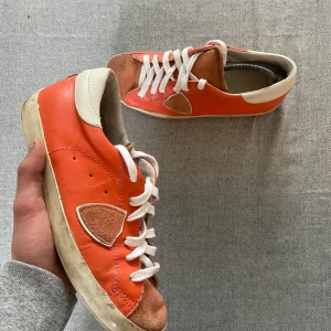 Orange sneakers med mockadetaljer från Philippe models  - Säljer ett par orange sneakers med vita snören och beige mockadetaljer på tå och sidan. Skorna har vit sula och häl, samt rund tå och platt sula. Perfekta för dig som vill sticka ut med färg på fötterna!