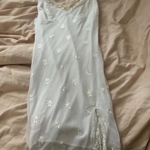 Vit blommig spetsklänning med slits - Söt vit kortklänning med broderade blommor och spetsdetaljer vid halsringning och slits. Klänningen har smala axelband och v-ringning.