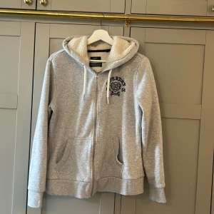 Grå hoodie med dragkedja från Superdry - Mysig grå hoodie från Superdry med dragkedja och broderad logga på bröstet. Fodrad med vit teddyfleece i huvan för extra värme. Två fickor framtill och justerbar huva med snören. Väldigt bra skick 