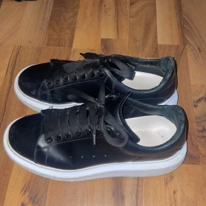 Svarta sneakers från Alexander McQueen - Säljer ett par svarta sneakers från Alexander McQueen med vit, tjock sula och svarta skosnören. Skorna har rund tå och är tillverkade i skinn. Klassisk och stilren design med logga i guld på hälen. Ingen låda kvar. 