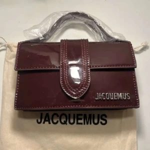 Vinröd handväska från Jacquemus - Säljer en snygg vinröd handväska från Jacquemus i glansigt material med silverfärgad logga. Väskan har ett stilrent och kompakt format med handtag och lockstängning. Axelremsband tillkommer. 💕