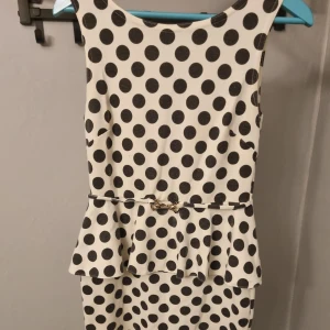 Vit och svart prickig klänning med volang - Säljer en vit klänning med stora svarta prickar och volang i midjan. Klänningen är ärmlös och har en markerad midja med ett smalt bälte och dragkedja i ryggen. Perfekt för dig som gillar retrostil!