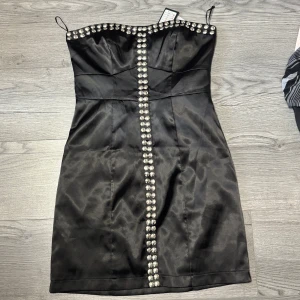Svart kort klänning från Vila - Säljer en svart axelbandslös ( det finns axelband i  påse på sista bilden) kort klänning från Vila med dekorativa strassknappar längs bysten och framtill. Klänningen har figurnära passform och är perfekt för festliga tillfällen. Materialet är glansigt och känns lyxigt.