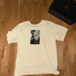 Tupac T-shirt - Säljer en beige T-shirt från Pull&Bearmed Tupac-tryck på bröstet och texten 'TUPAC' på ryggen. T-shirten har använts 1-2 gånger och färgen är vit/gul. Väldigt skön att ha på sig och passar väldigt bra till det mesta! Storleken är Medium (M)