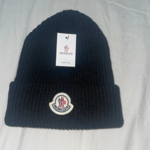 Svart ribbad mössa från Moncler - Svart ribbstickad mössa från Moncler med uppvikt kant och Moncler-logga framtill. Klassisk och stilren design som passar perfekt till kyliga dagar. Mössan är tillverkad i ett mjukt och värmande material. 