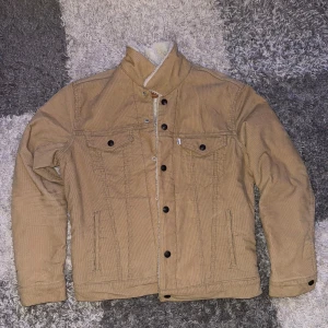 Beige sherpa-fodrad jeansjacka från Levi's - Säljer en beige jeansjacka från Levi's med mysigt vitt sherpa-foder. Jackan har klassisk krage, bröstfickor med knappar och knäppning framtill. Perfekt för kyligare dagar och riktigt snygg till jeans eller chinos.