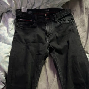 Svarta jeans från Tommy Hilfiger Layton Stretch - Säljer ett par svarta Tommy Hilfiger Layton Stretch jeans med extra slim fit. Byxorna har klassisk femficksdesign, röda sömdetaljer och diskret logga på bakfickan. Perfekta för dig som gillar en tajtare passform och stilren look.