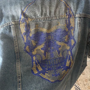 Oversized jeansjacka med döskalletryck - Säljer en oversized jeansjacka i blå denim. Jackan har slitna detaljer, knappar framtill och ett stort döskalletryck i guld och blått på ryggen. Perfekt statement-plagg för dig som vill sticka ut.