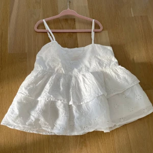 Vit broderad topp från Gina Tricot - Söt vit topp från Gina Tricot med smala axelband och volang. Toppen har broderade blommor och är perfekt för varma dagar. Lätt och luftig modell som ger en somrig känsla.