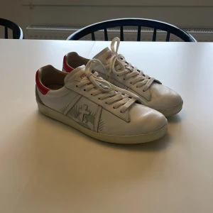 Vita premiata sneakers med röd häl och korsdetalj - Säljer ett par vita premiata sneakers med röd häl och vit snörning. Sidan har en broderad korsdetalj i grått och vitt. Skorna har platt sula och rund tå, perfekta för en avslappnad stil. De är  knappt använda och är i sjukt bra skick!😊
