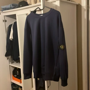 Mörkblå sweatshirt från Stone Island - Säljer en mörkblå sweatshirt från Stone Island med klassisk patch på ärmen. Tröjan har rund halsringning och långa ärmar med breda muddar. Perfekt för en stilren och avslappnad look.köpte den av min kompis då den inte passade han.