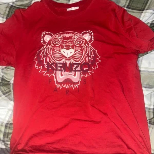 Röd t-shirt med tigertryck från Kenzo - Säljer en röd t-shirt från Kenzo med ikoniskt tigertryck och texten 'Kenzo Paris'. Köptes för 1200kr, ganska använd men inga märken på eller liknande. Använt mycket men bra skick 
