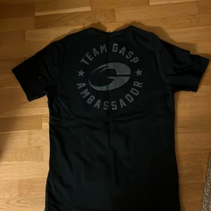 Svart t-shirt från GASP med tryck - Säljer en svart t-shirt från GASP i storlek XL. T-shirten har tryck med texten 'TEAM GASP AMBASSADOR' både på bröstet och ryggen. Perfekt för dig som gillar träning och vill ha en stilren look.