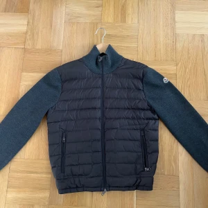 Grå cardigan jacka från Moncler - Säljer en stilren svart och mörkgrå cardigan jacka från Moncler. Jackan har quiltad framsida i dun och stickade ärmar samt rygg. Hög krage, dragkedja framtill och två fickor med dragkedja. Perfekt för lager-på-lager och snygg till vardags. Passar M/L